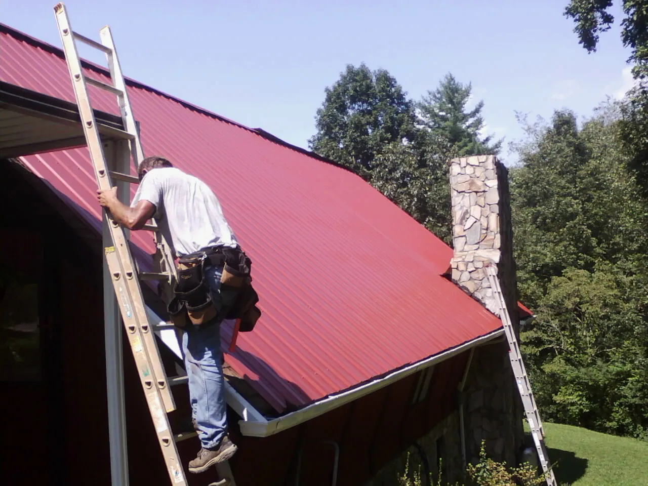 Roofer on a ladder beside a red metal roof and stone chimney — Soffit & Fascia Repair by Roofing Brothers in Grand Haven